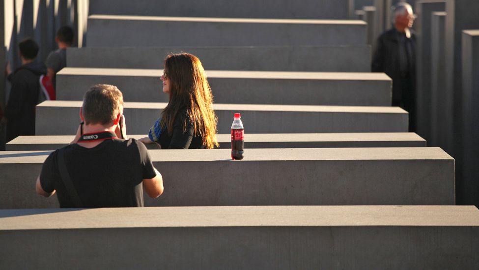 Zwei junge Touristen stehen am 19.10.2014 zwischen Steelen im Holocaust-Mahnmal in Berlin und machen Fotos. Der Mann fotografiert die Frau, dabei haben sie eine Cola-Flasche auf einer Steele abgestellt. (Quelle: dpa/Wolfram Steinberg)