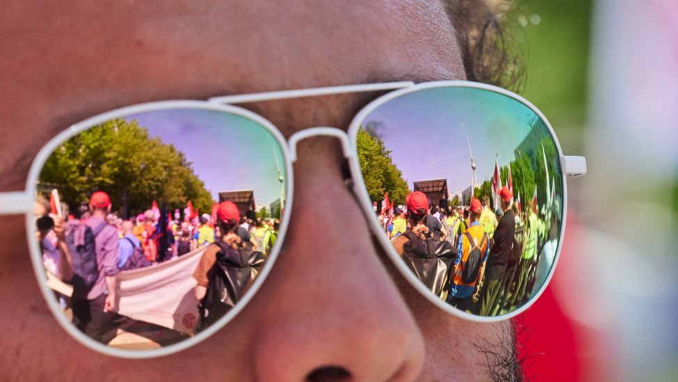 01.05.2025, Berlin: Auf einer Spiegelbrille sieht man, wie sich Teilnehmer einer Demonstration zum 1. Mai auf der Karl-Marx-Alle versammeln.(Quelle:dpa/J.Carstensen)